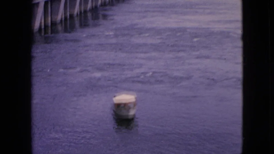 The small covered boat sits farther across the dark, purple-tinged river surface, with partial dock or bridge pilings along the upper edge of the frame.