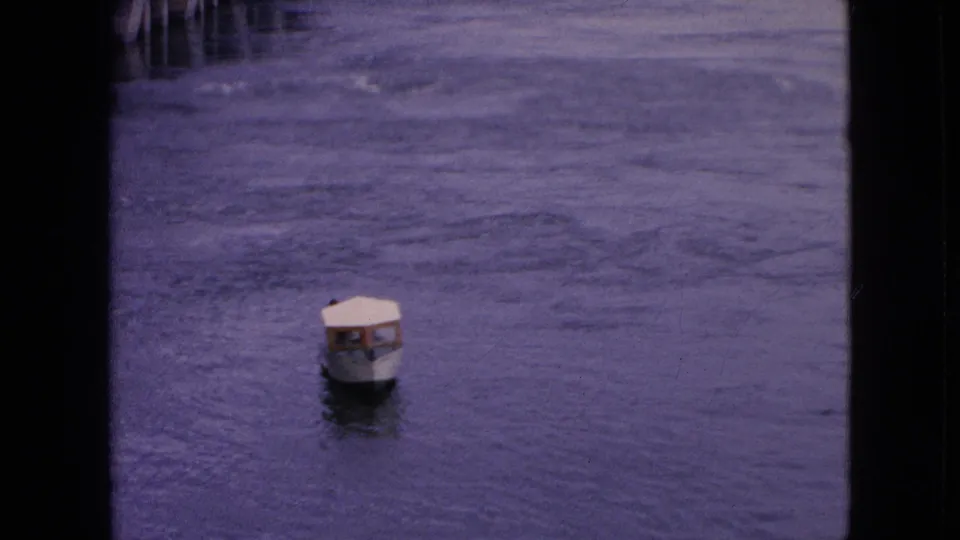 A distant covered motorboat appears near the lower portion of the frame, surrounded by broad rippled water in soft, grainy 8mm color.