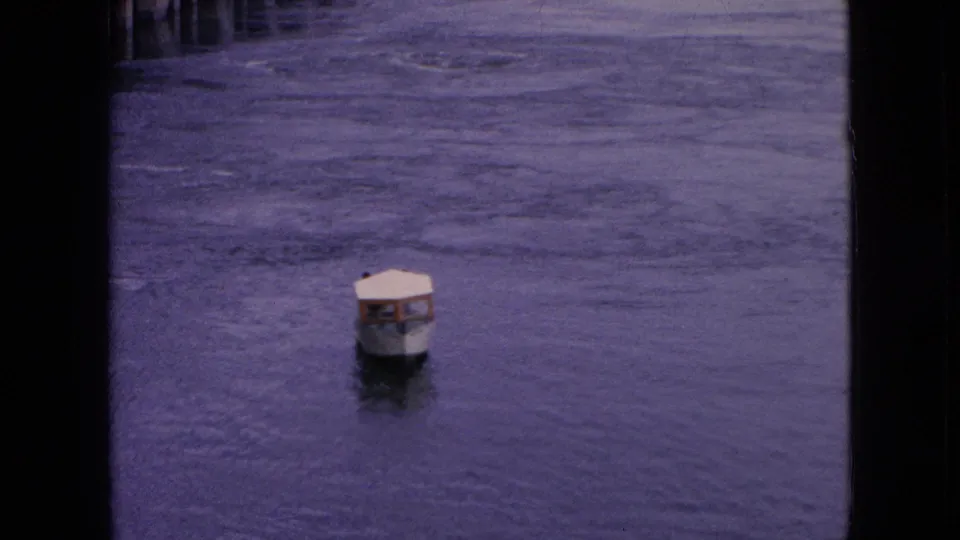 The covered boat is framed near center against textured river water, with indistinct occupants visible beneath the canopy and dark 8mm frame borders at the sides.