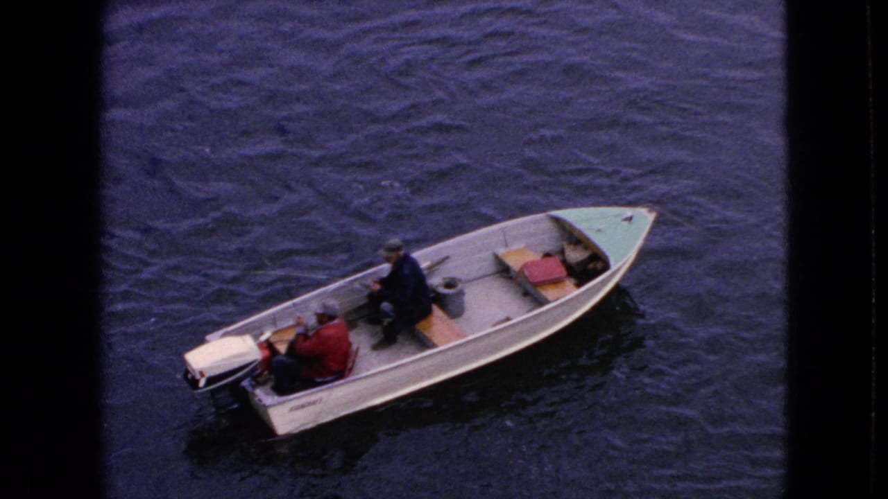 1964 home-movie frame showing two adult men in a small light-colored motorboat, with one in a red jacket near the outboard motor and another in a dark jacket seated forward on the water near Niagara Falls, New York.