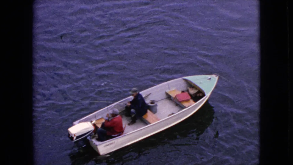 1964 home-movie still of a small aluminum motorboat angled across the frame, with two adult men seated inside and dark rippling water filling the background.