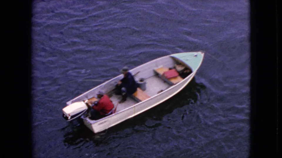 Final 1964 8mm frame showing two adult men in a small motorized boat, the stern outboard visible at left and the green bow pointing across dark rippling water.