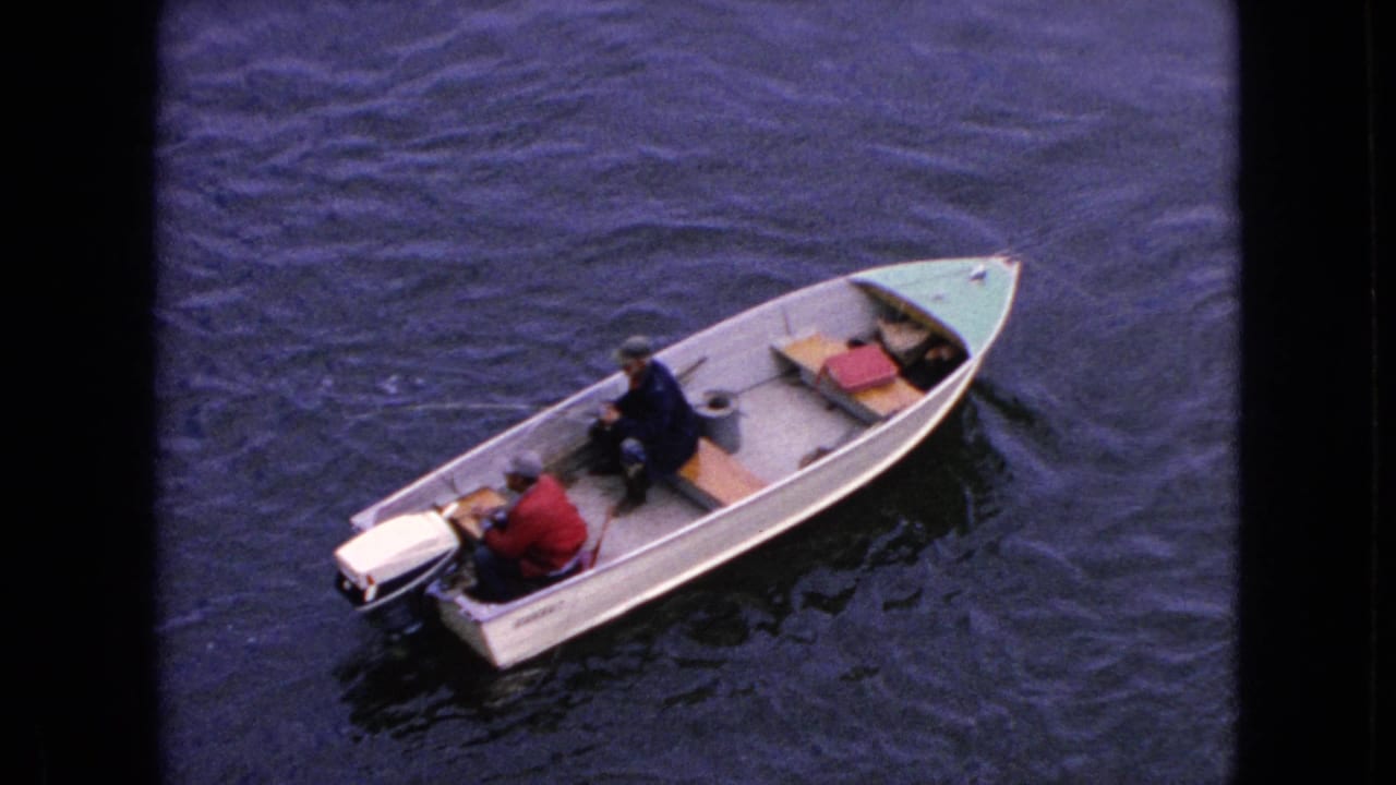 High-angle 1964 home-movie view of two adult men in an aluminum motorboat on the water near Niagara Falls, New York, with a white outboard motor at the stern.