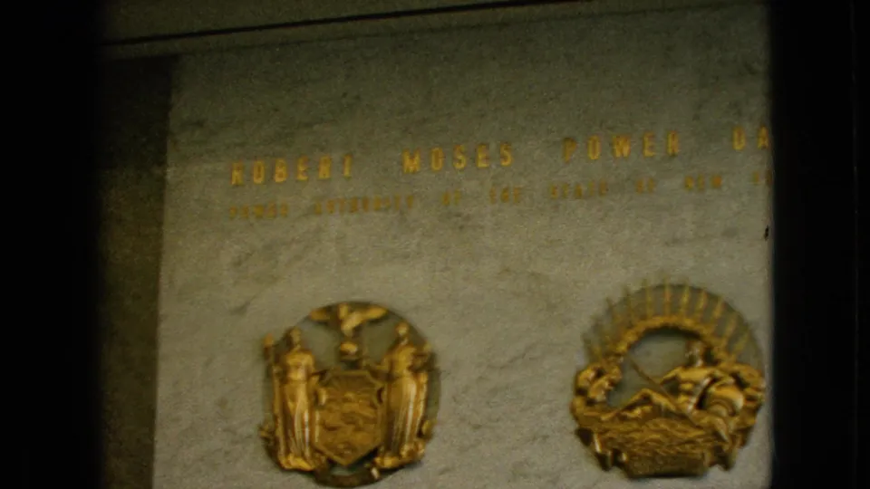 Close view of a stone plaque reading Robert Moses Power Dam, with gold lettering and two decorative emblems visible in a 1964 home-movie frame.