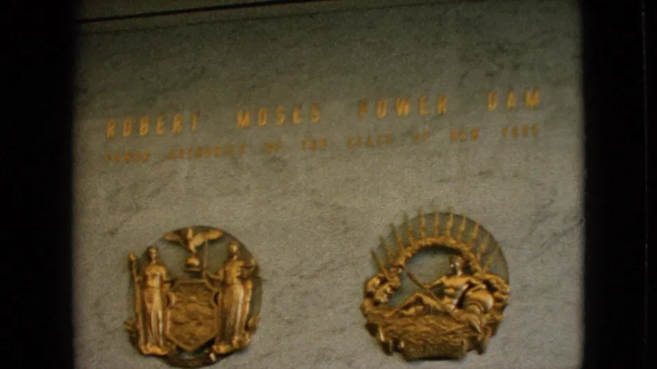 Home-movie close-up of the Robert Moses Power Dam plaque, showing gold letters across stone and two raised decorative emblems below.