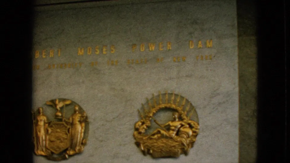 Close 8mm frame of the Robert Moses Power Dam plaque, with the name partly visible above two gold-toned emblems on a stone wall.