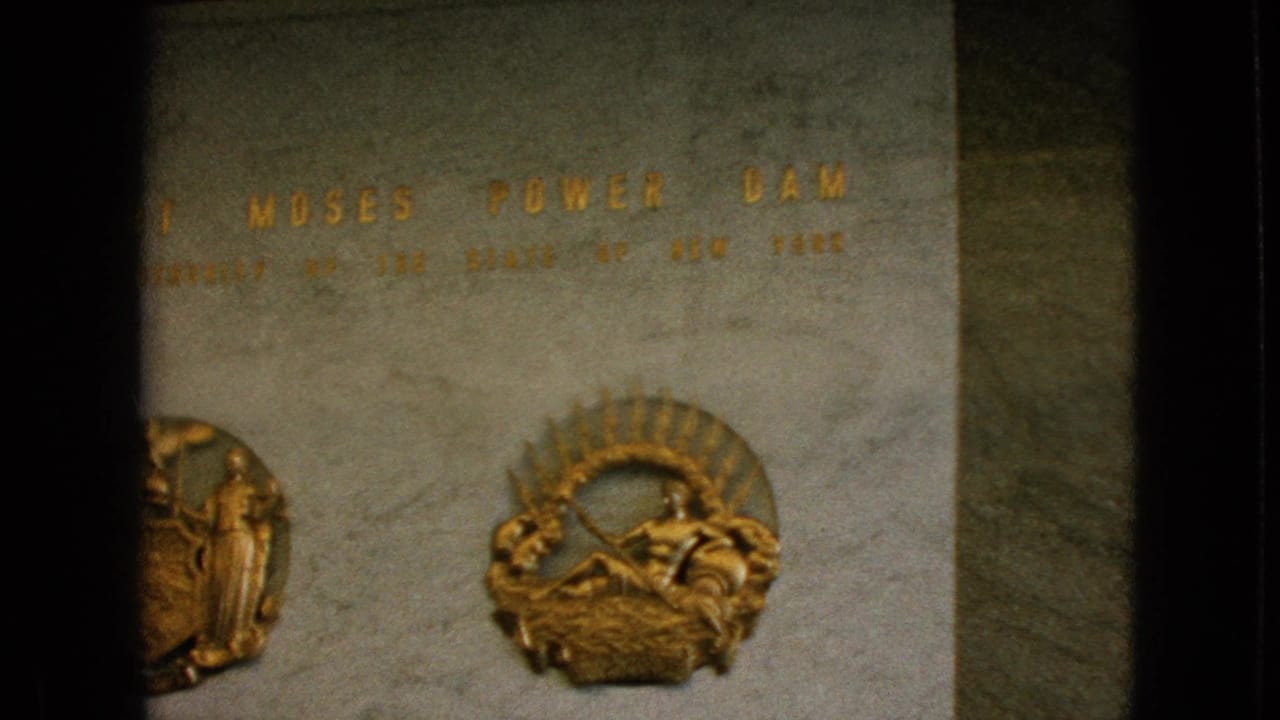1964 Niagara Falls home-movie frame showing part of the Robert Moses Power Dam inscription on polished marble with raised gold lettering and emblem reliefs.