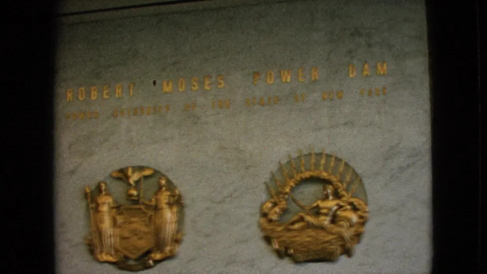 The frame widens to show the Robert Moses Power Dam inscription across polished stone, with the New York state seal relief and a sunburst-style emblem below.