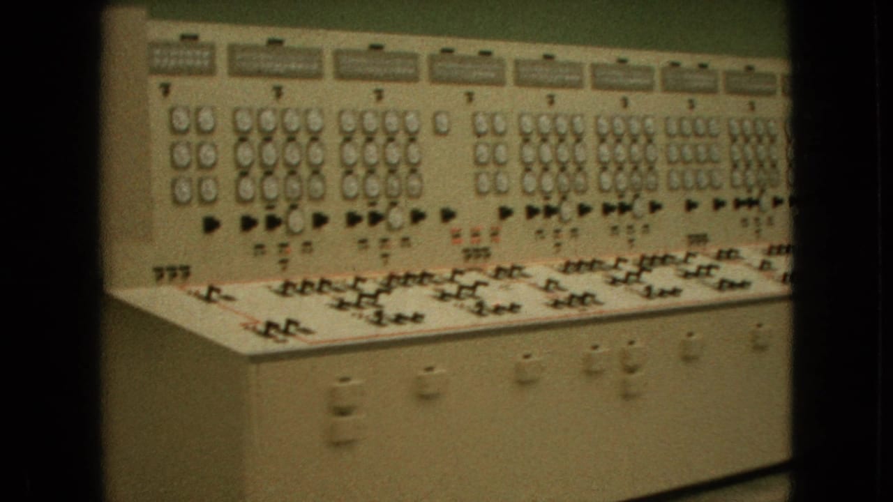 1964 United States 8mm frame showing a long beige analog control panel with clustered gauges, switches, labels, and indicator lights along a green wall.