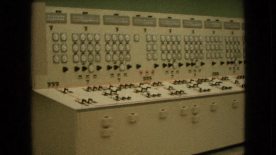 1964 United States 8mm frame showing a long beige analog control panel with clustered gauges, switches, labels, and indicator lights along a green wall.