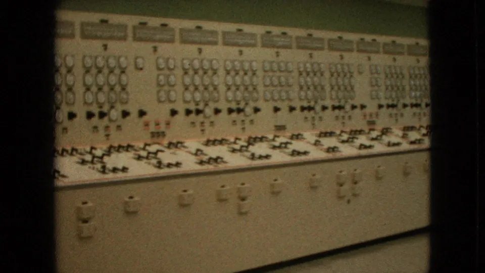 1964 United States 8mm frame of a wall-length beige control console with repeated dials, switches, and illuminated sections, bordered by dark film-frame spacing.