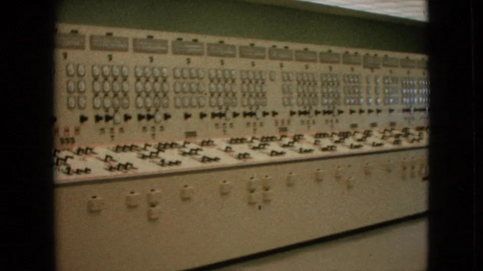 1964 United States 8mm frame showing the long control panel from a slightly lower angle, with dense rows of analog instruments, switches, and cabinet doors below.
