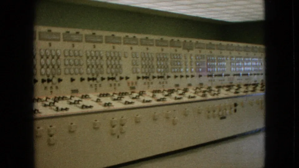 1964 United States 8mm frame showing the extended beige control panel continuing along the wall, with repeated gauges, switches, lighted indicators, and a bright ceiling strip.