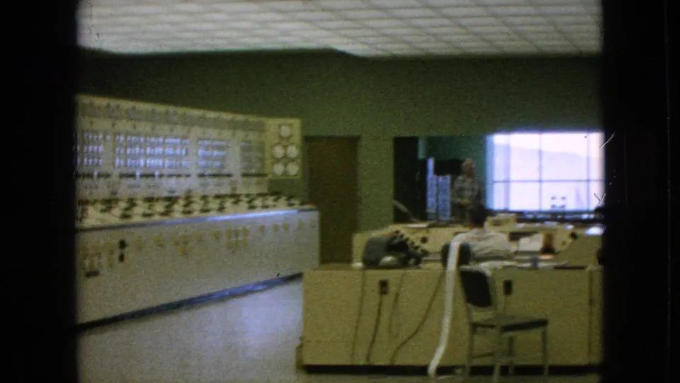 1964 Niagara Falls, New York footage showing the control console and a person seated at a desk near a bright window inside the industrial room.