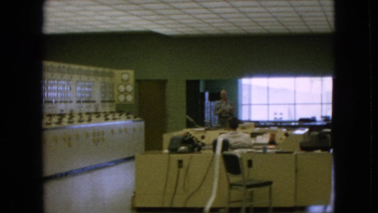 1964 8mm frame from Niagara Falls, New York metadata showing a room-sized control panel, desk area, seated worker, standing man, cords, chairs, and a bright window.
