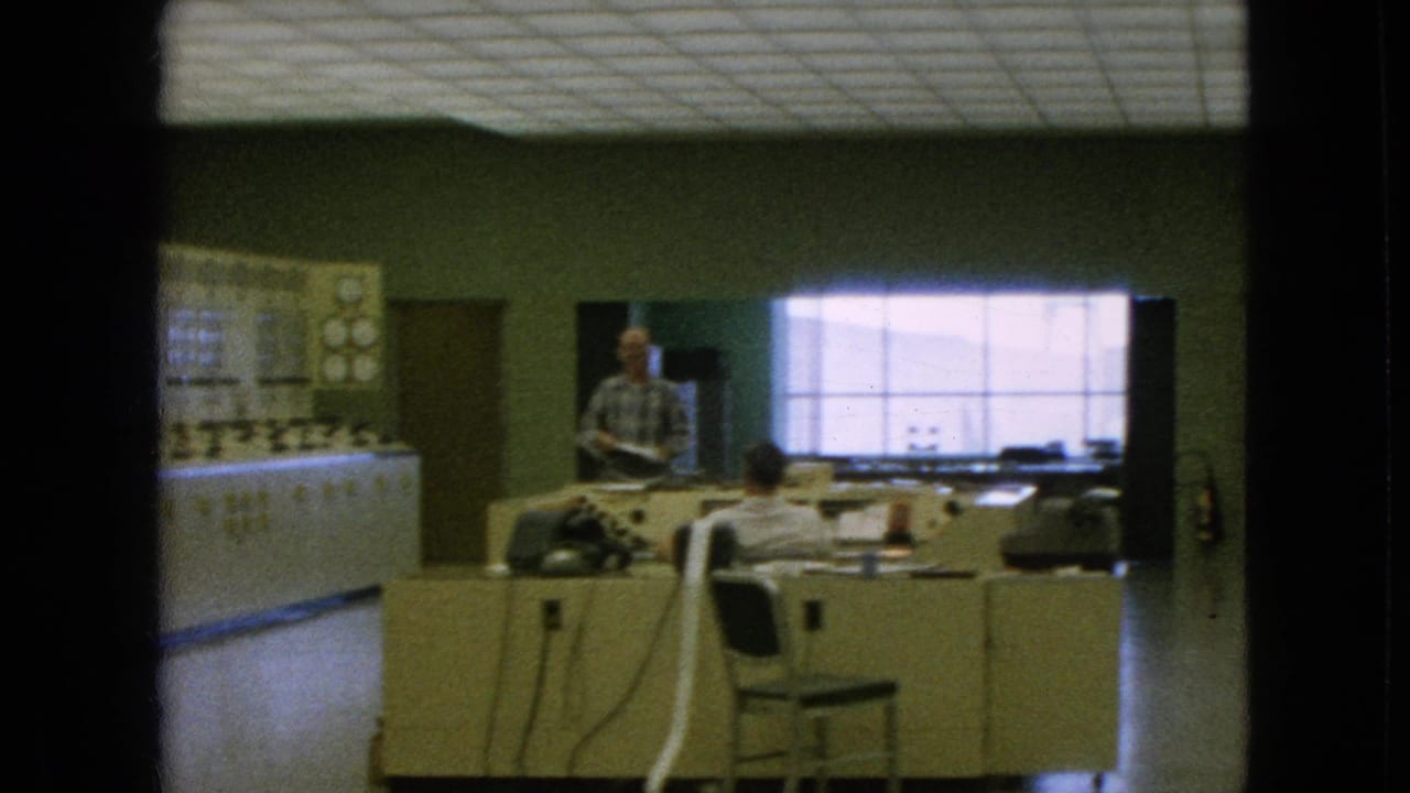 A standing man and seated worker appear beside desks and a large wall control panel.