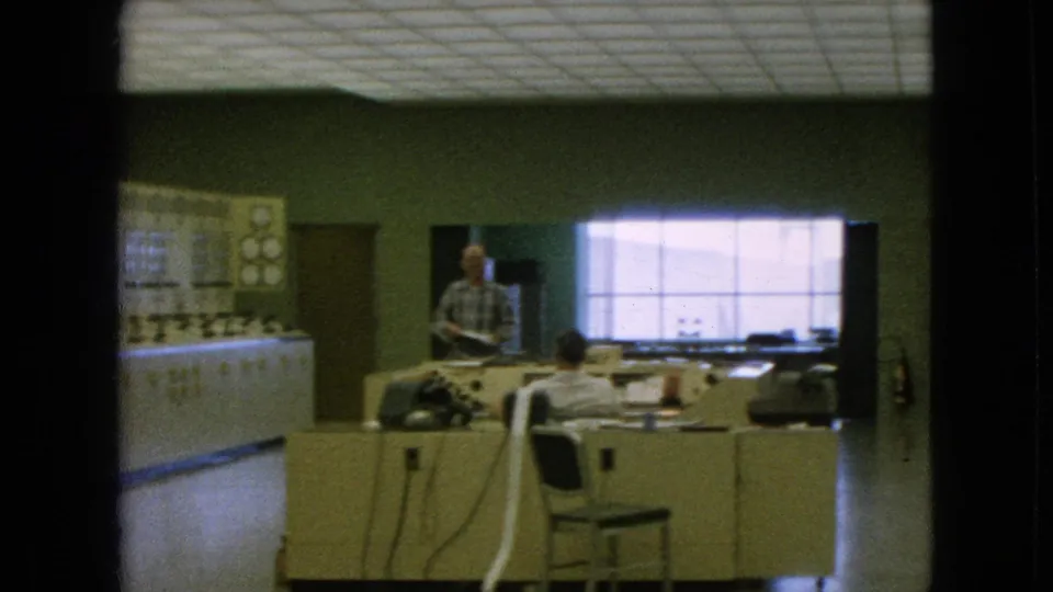1964 Niagara Falls, New York metadata frame showing a man standing near a desk while another worker sits facing equipment beside a long analog control panel.