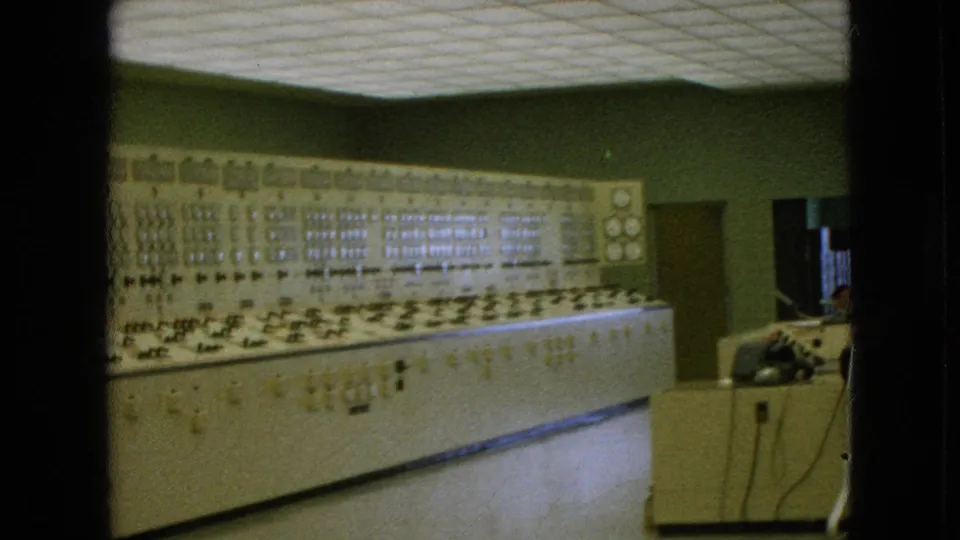 1964 8mm frame, based on supplied metadata, showing the control-room panel more prominently with rows of gauges, switches, a desk corner, cords, and a chair.