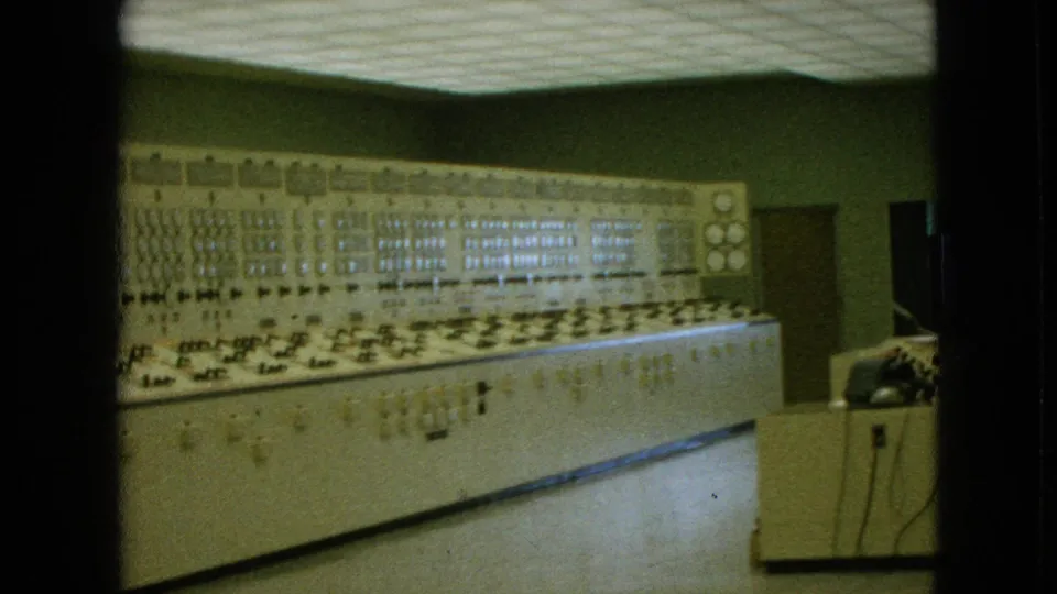 1964 Niagara Falls, New York metadata frame showing a long analog control panel with many dials and switches, overhead ceiling panels, and desk equipment at the right edge.