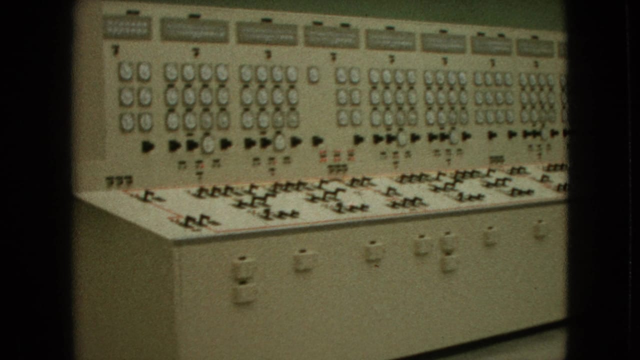 1964 8mm frame showing a hydroelectric-style electrical control panel with analog gauges, toggle switches, indicator lamps, and labeled sections.