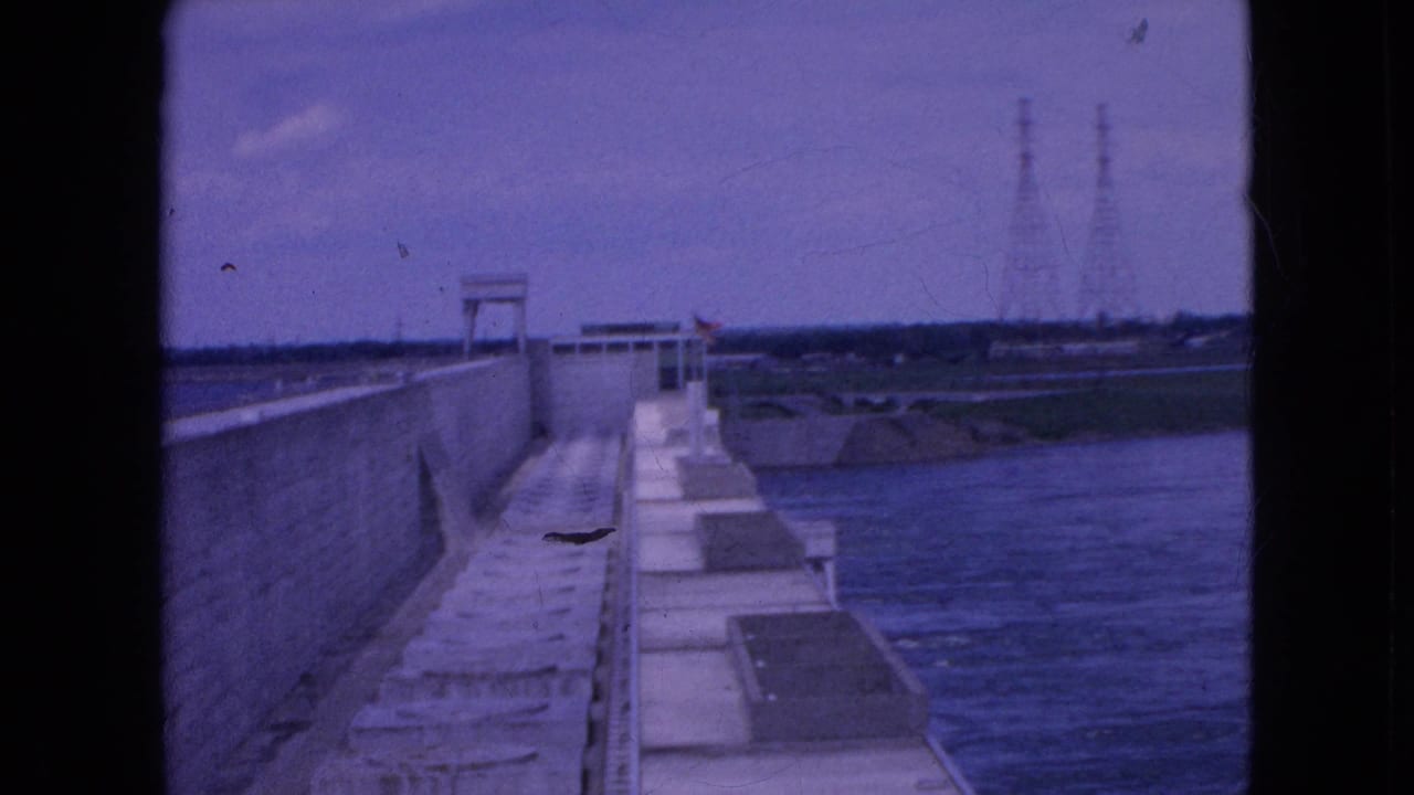 Concrete dam walkway beside held water with transmission towers in the distance.