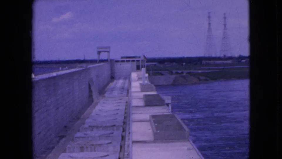 1964 8mm exterior frame of a concrete dam wall and walkway beside water, with utility towers visible beyond the structure.