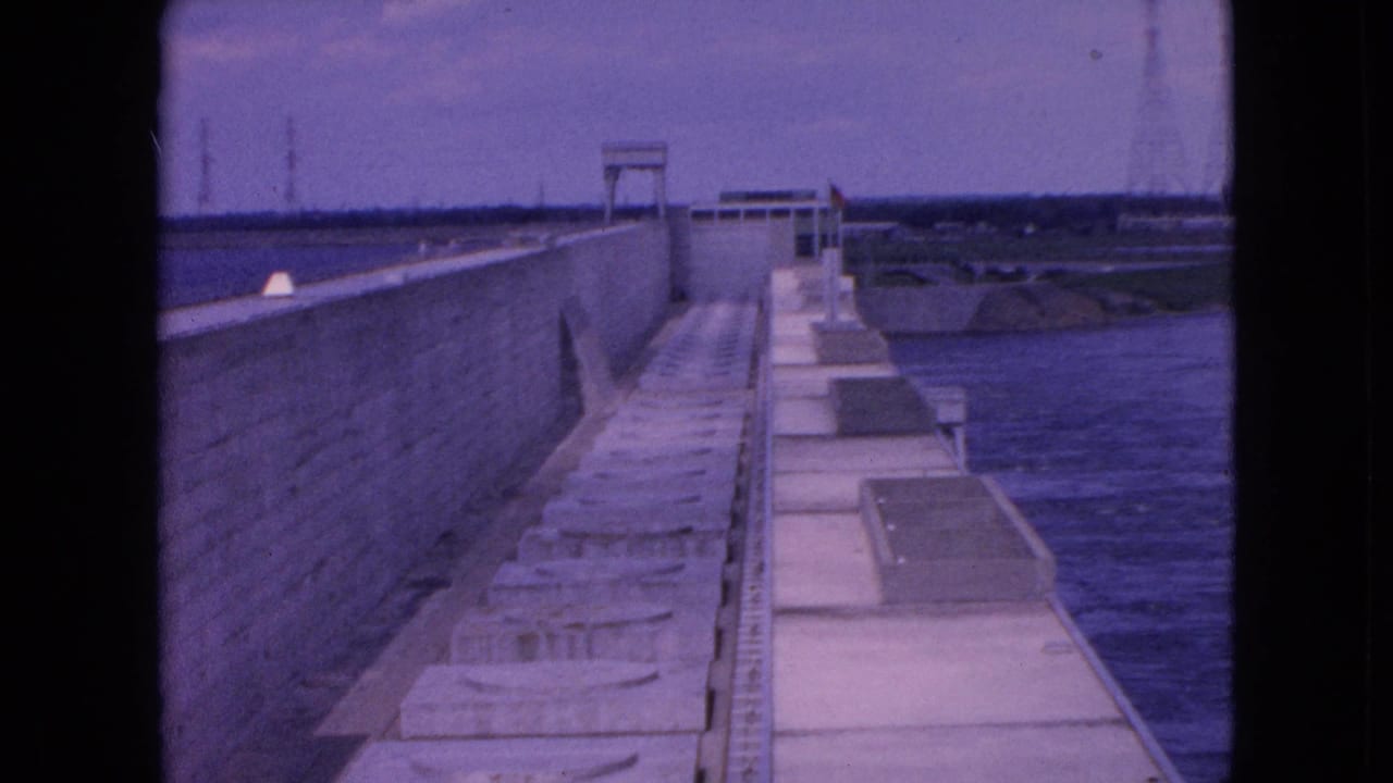 1964 8mm frame from Niagara Falls, New York, showing a long concrete industrial walkway between a high wall and a waterway.