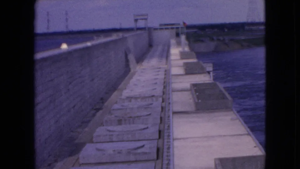 1964 Niagara Falls frame showing a wider view of the concrete causeway or bridge work, with open water to the right and distant utility towers under a dim sky.