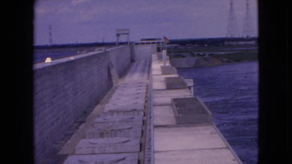 1964 Niagara Falls home-movie frame looking along the concrete structure beside water, with distant buildings, utility lines, and a faintly lit sky in the background.