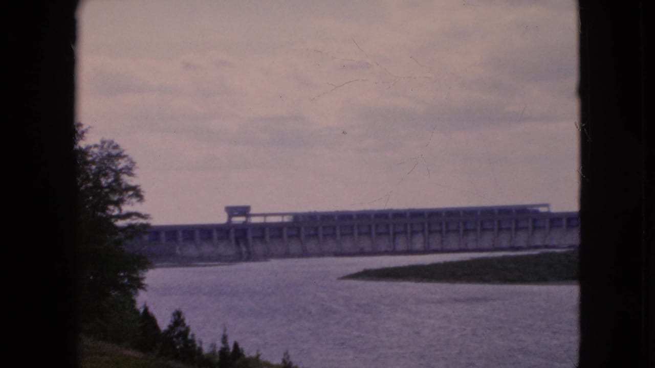 1964 8mm home-movie frame from the Niagara Falls, New York area showing a long concrete dam across a wide river under an overcast dusk sky, with dark foreground silhouettes at the sides.