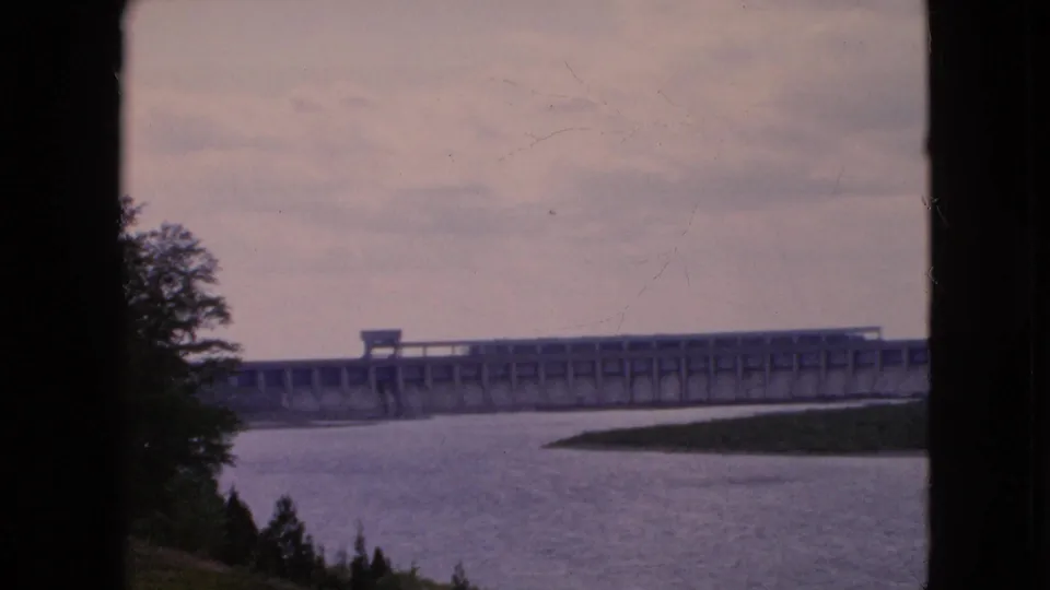 1964 8mm home-movie frame from the Niagara Falls, New York area showing a long concrete dam across a wide river under an overcast dusk sky, with dark foreground silhouettes at the sides.