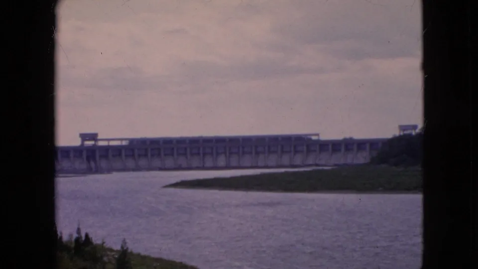1964 Niagara Falls-area 8mm frame showing the dam extending across the river, a low grassy bank in front, and dark structural silhouettes framing the view.