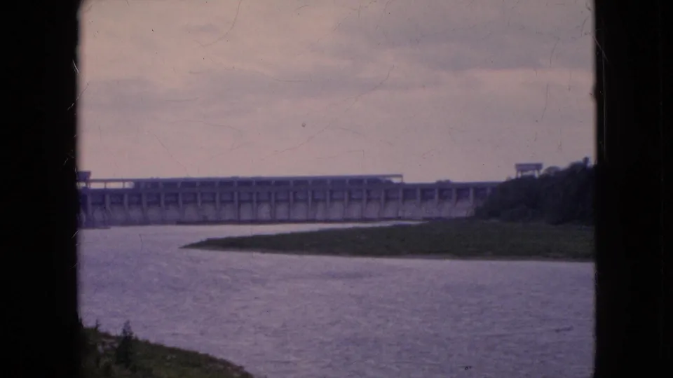 Final 1964 8mm frame of the Niagara Falls-area river dam, showing the concrete structure in the distance, calm water in front, and silhouetted foreground edges from the filming position.