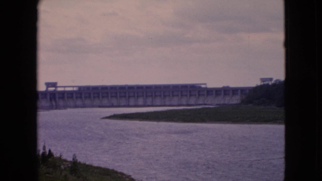 1964 8mm home-movie frame from Niagara Falls, New York, showing a broad concrete dam beyond calm river water and a grassy bank, seen through dark foreground edges.