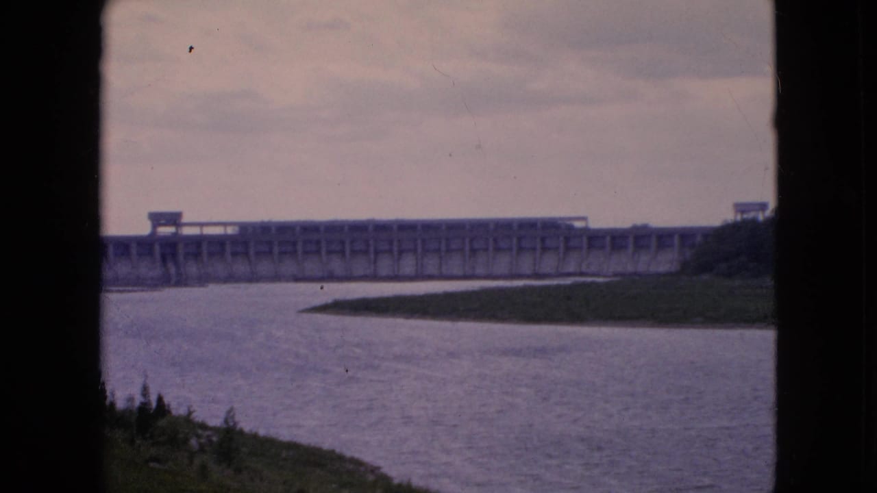 Wide 1964 Niagara Falls river view with a concrete dam on the horizon, muted sky above, and a dark vertical edge dividing the home-movie composition.