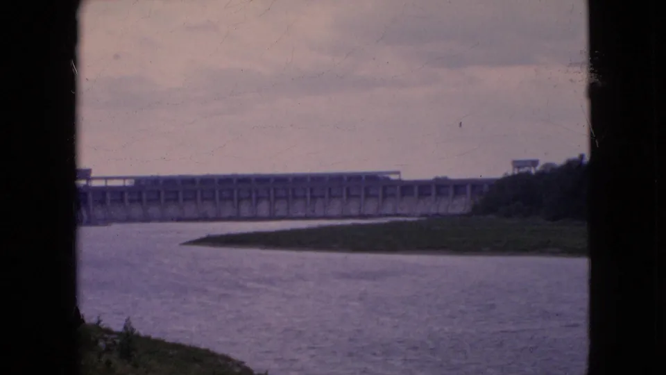 1964 8mm frame showing a long concrete dam across the river at Niagara Falls, New York, with a narrow grassy bank and dark film-frame edges in the foreground.