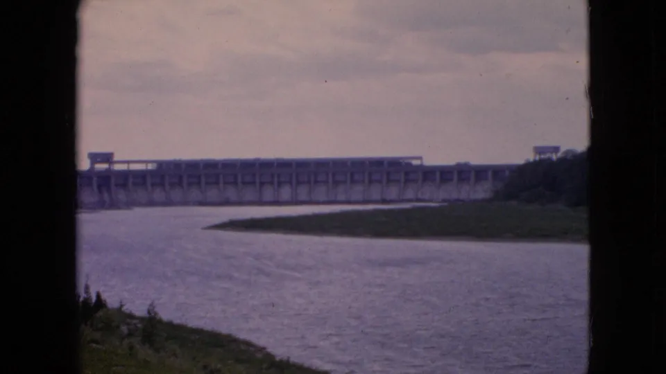 Final 1964 Niagara Falls home-movie frame with calm river water, a grassy point, and a large concrete dam stretching across the background beneath a muted sky.