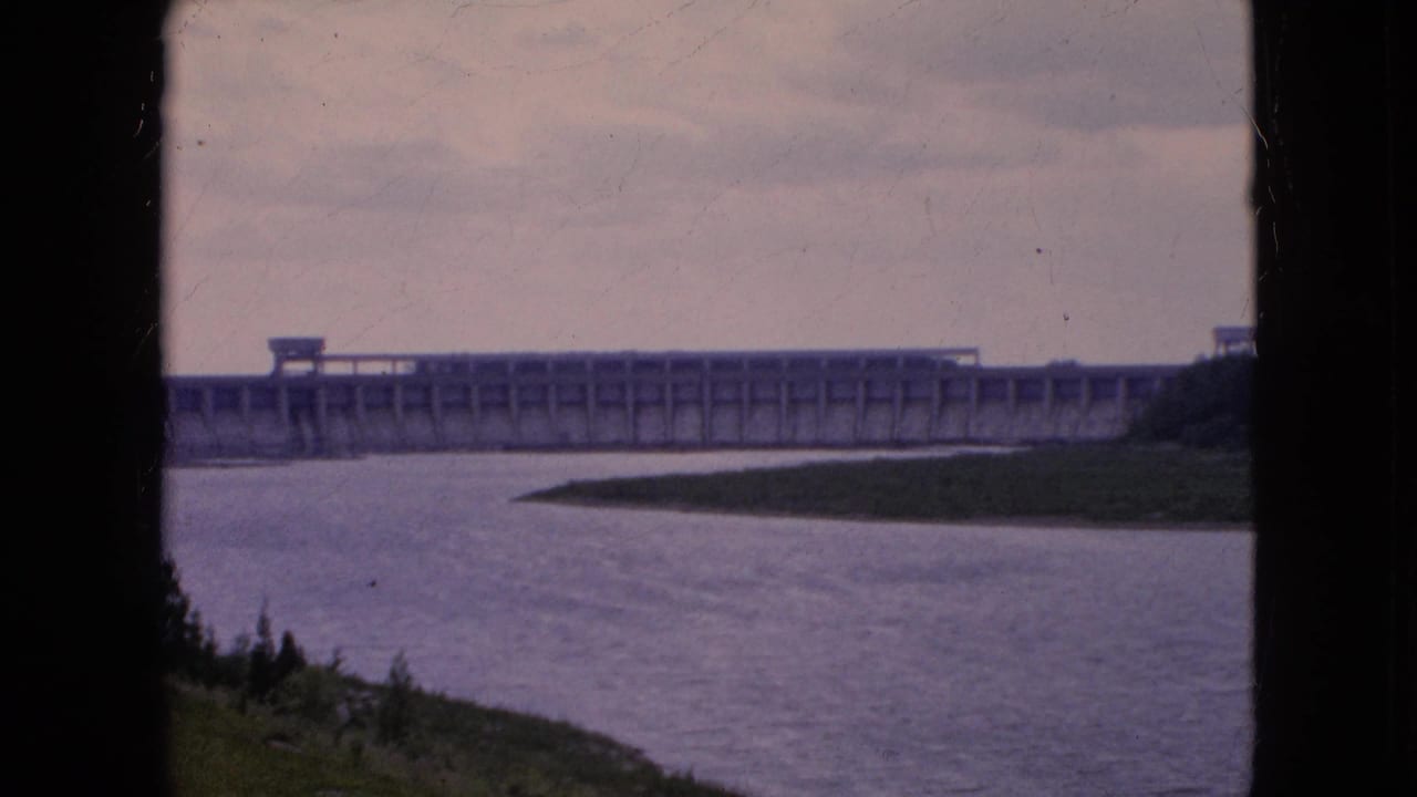 1964 8mm frame showing a distant concrete dam spanning a wide river in the Niagara Falls, New York area, with dark frame edges and a grassy foreground bank.