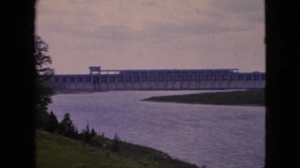 1964 8mm frame of a distant concrete dam behind a broad river, with trees at the left edge and a grassy foreground slope.