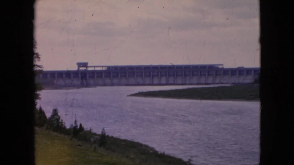 1964 8mm frame showing a wide river bending toward a distant concrete dam in the Niagara Falls, New York area, with shoreline vegetation and visible film grain.