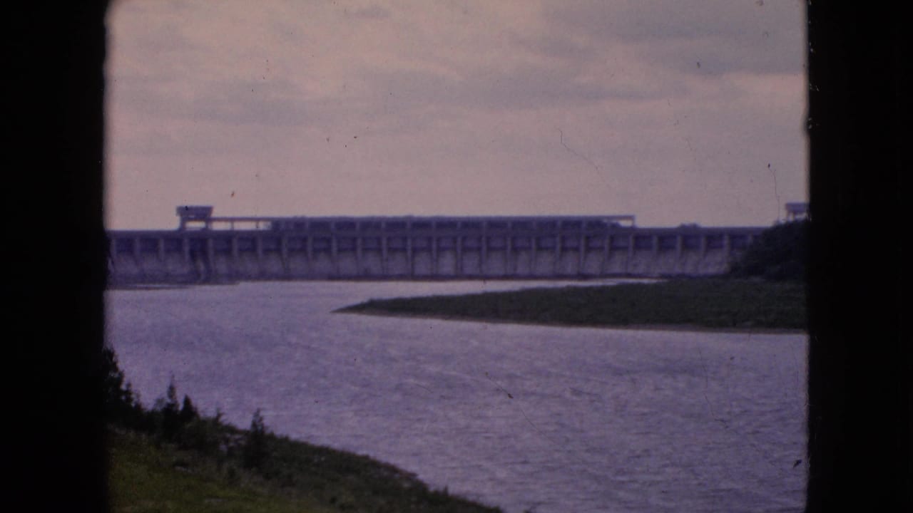 1964 8mm frame showing a distant concrete dam spanning a broad river, with a grassy bank in the foreground and an overcast sky above.