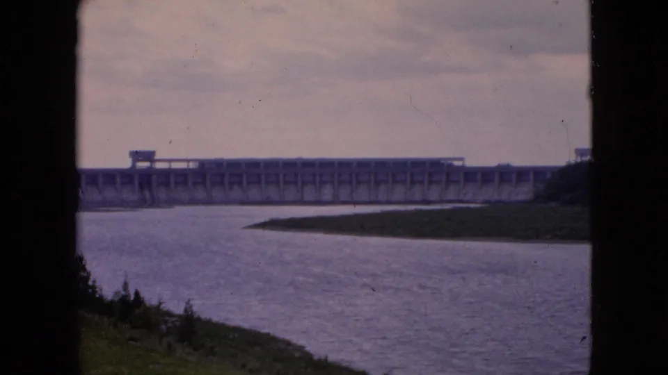1964 8mm frame showing a distant concrete dam spanning a broad river, with a grassy bank in the foreground and an overcast sky above.