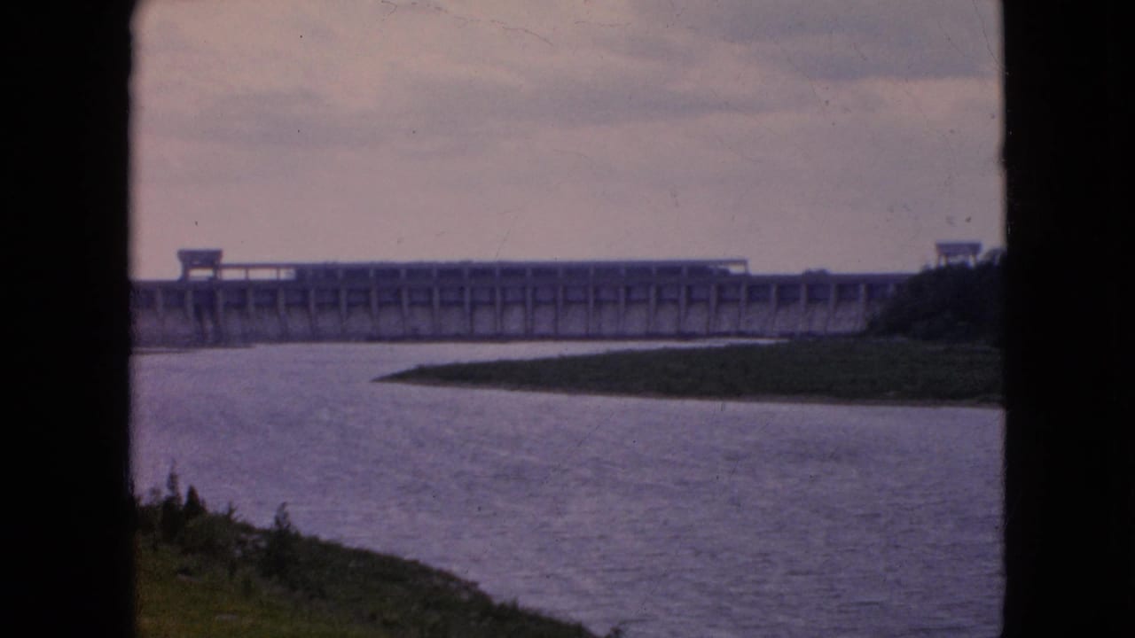 Distant view of a long concrete dam over a calm river, framed by dark 8mm gate edges and soft faded color.
