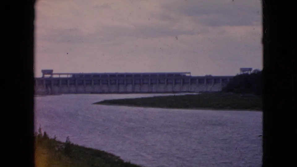 8mm home-movie frame with a broad river in the foreground, a distant dam along the horizon, and dark film-frame borders at the sides.