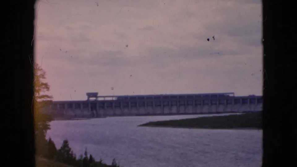 Final 1964 8mm frame showing the dam from a distance across the river, with cloudy sky and small dark specks overhead that may be aircraft or film debris.