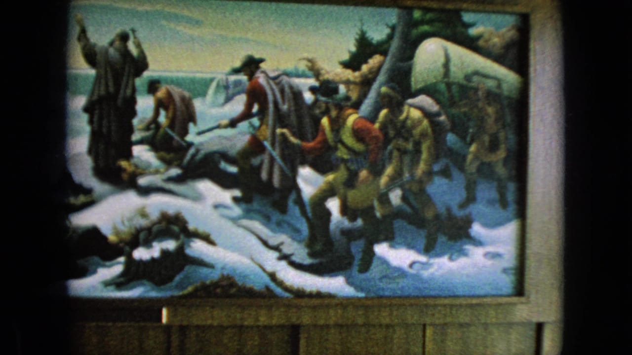 1964 8mm frame showing a painted snowy expedition or soldier diorama, with bundled figures gathered around a long boat inside a dark framed viewing area.
