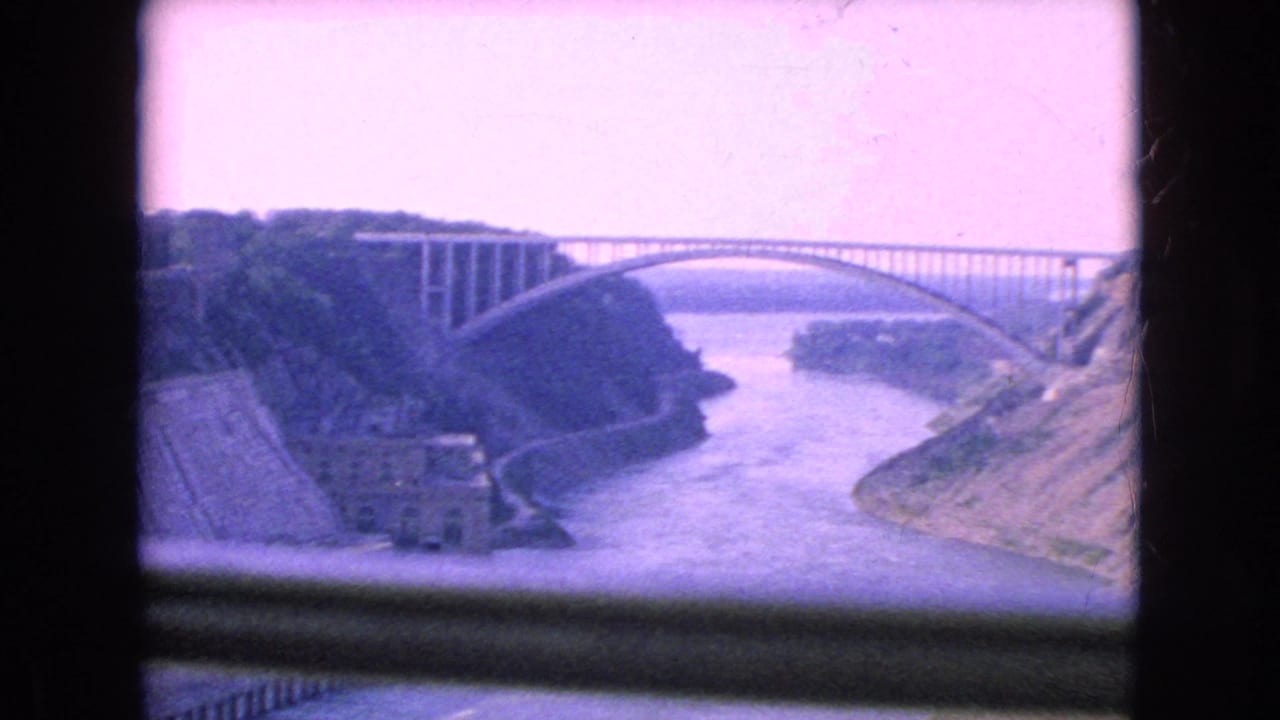 1964 8mm frame of a large arched bridge over a river gorge, viewed through a dark rectangular frame with a purple film cast.