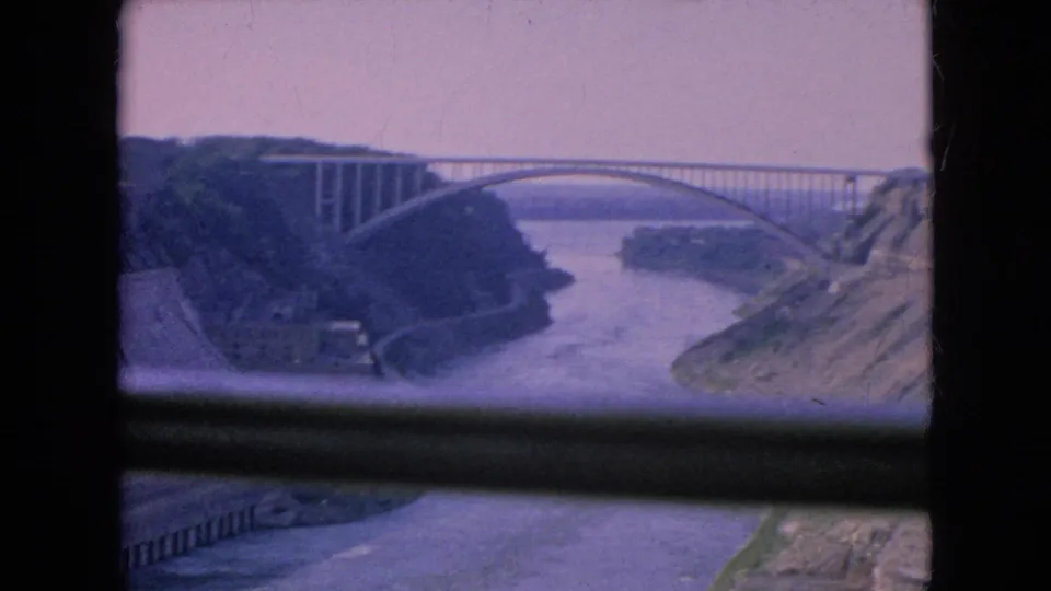 1964 8mm frame of an arched river bridge with wooded and rocky gorge banks, softened by grain and magenta color shift.