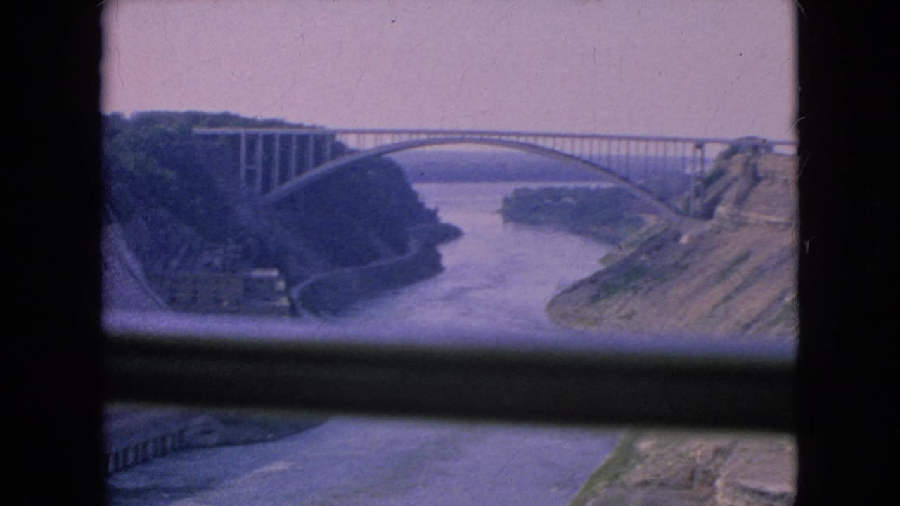 1964 Niagara Falls, New York home-movie frame showing a wide river flowing through a rocky gorge beneath a large arched bridge, viewed through a dark foreground window frame.