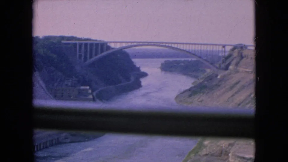 1964 Niagara Falls, New York home-movie frame showing a wide river flowing through a rocky gorge beneath a large arched bridge, viewed through a dark foreground window frame.
